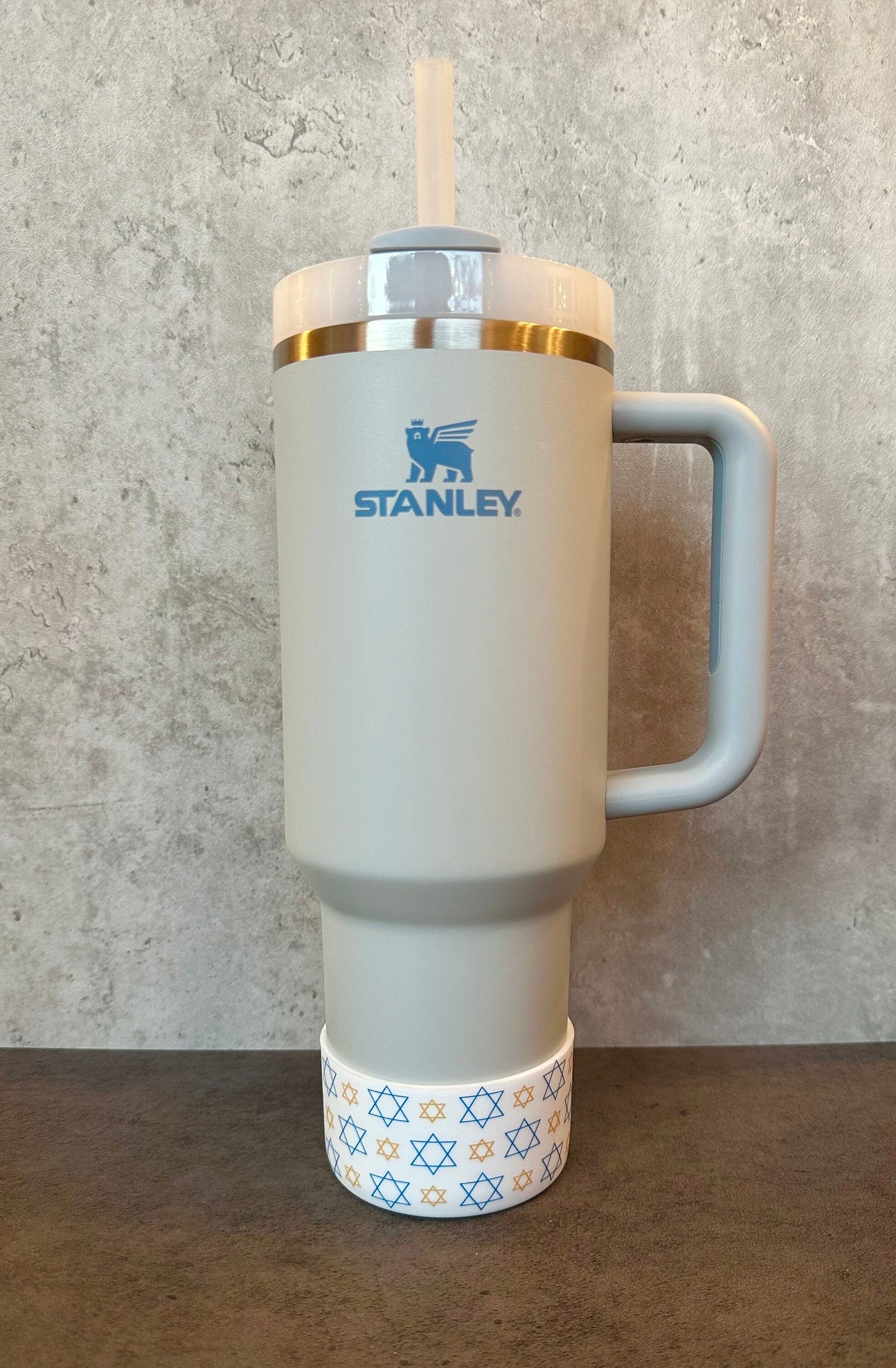 May include: A gray stainless steel travel mug with a blue bear logo that says 'STANLEY.' The mug has a gray handle and a white lid with a straw. The bottom of the mug has a white band with a blue and gold star of David pattern.
