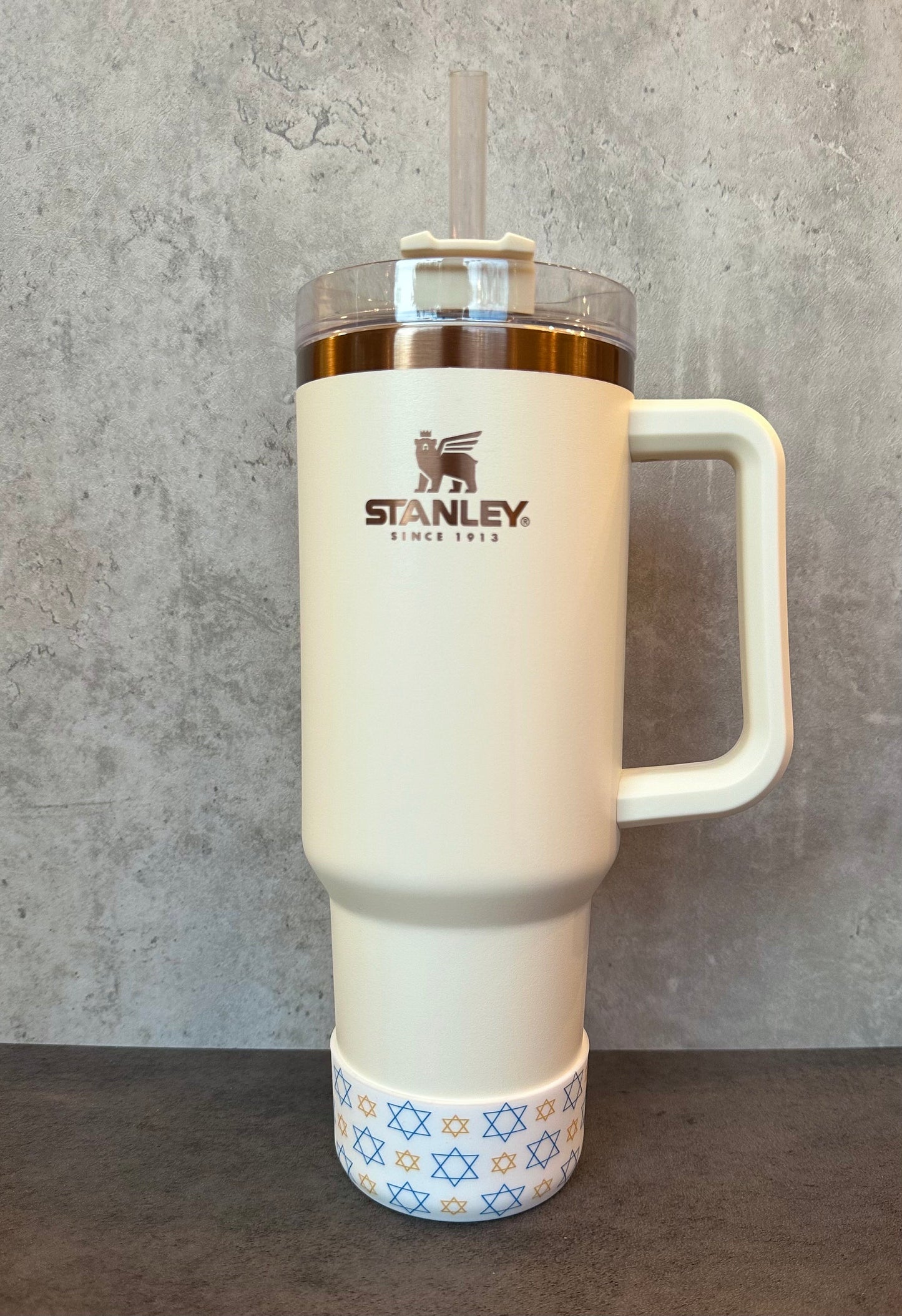 May include: A white stainless steel travel mug with a gold lid and handle. The mug has a gold logo that says 'STANLEY. SINCE 1913.' The bottom of the mug has a blue and gold star of David pattern.