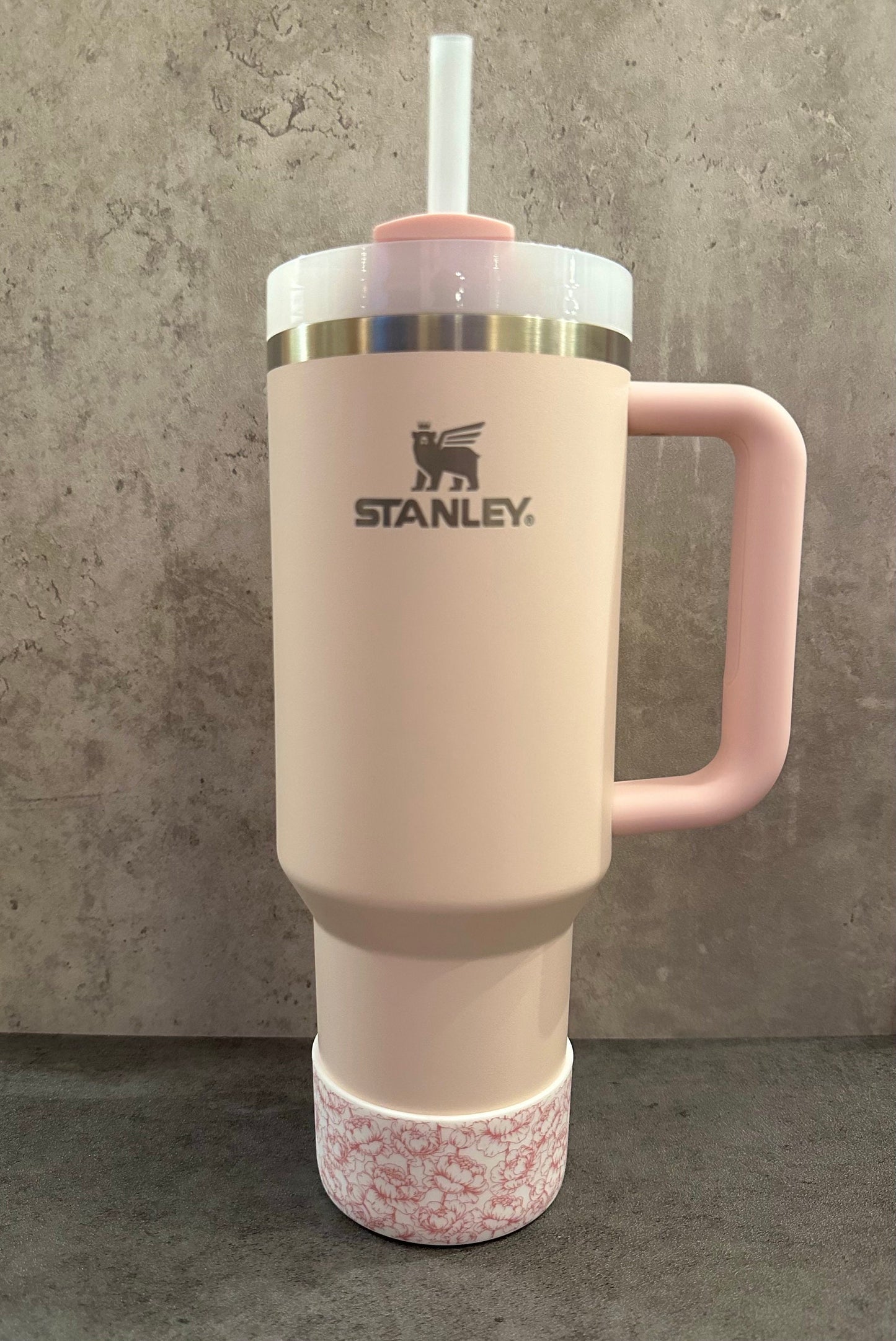 May include: A pink stainless steel travel mug with a handle and a straw. The mug has a floral pattern on the bottom and the Stanley logo on the side.