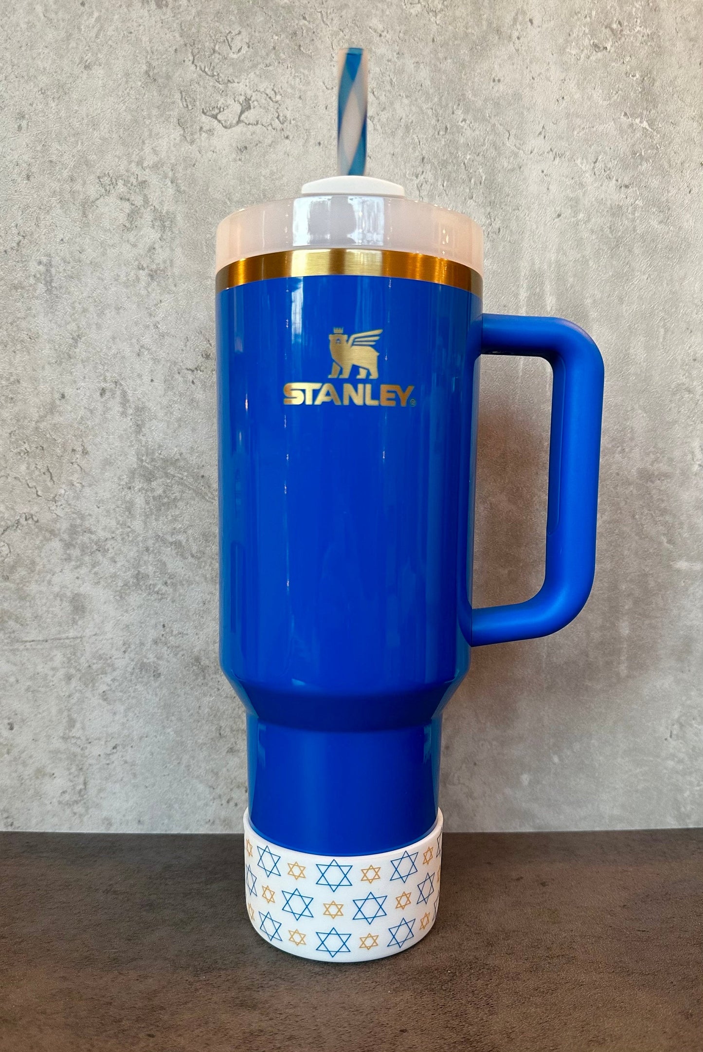 May include: A blue stainless steel travel mug with a gold lid and handle. The mug has a gold logo that says 'STANLEY.' The mug is sitting on a brown surface and has a white band with gold stars around the bottom.