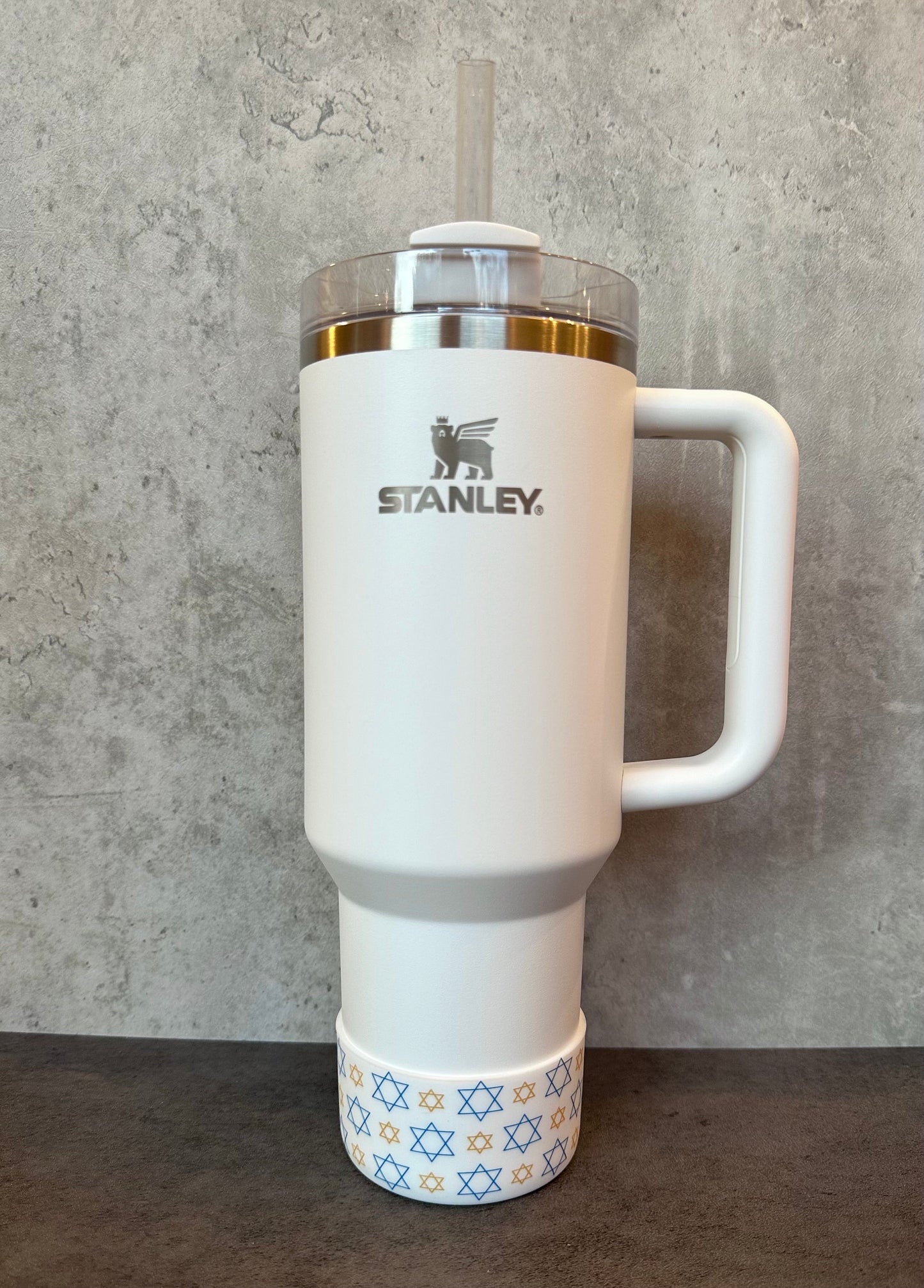 May include: White Stanley travel mug with a handle and a blue and gold star pattern around the bottom. The mug has a silver lid and a straw. The Stanley logo is printed on the mug.