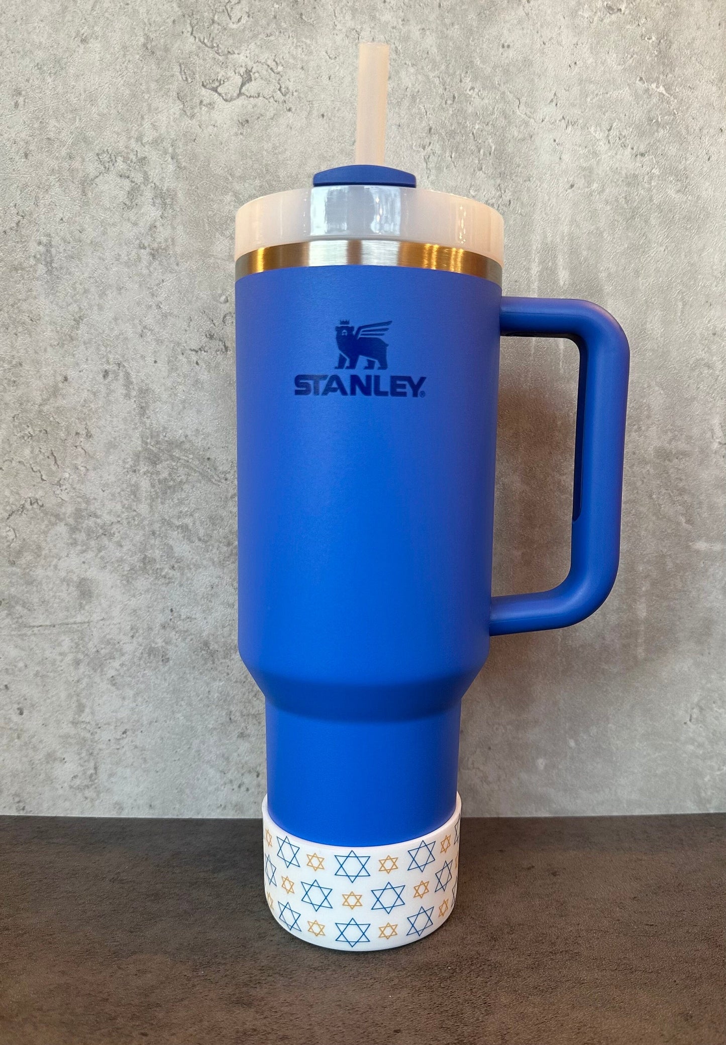 May include: A blue stainless steel travel mug with a handle and a straw lid. The mug has a white band with a pattern of gold stars of David. The mug has the Stanley logo on the side.