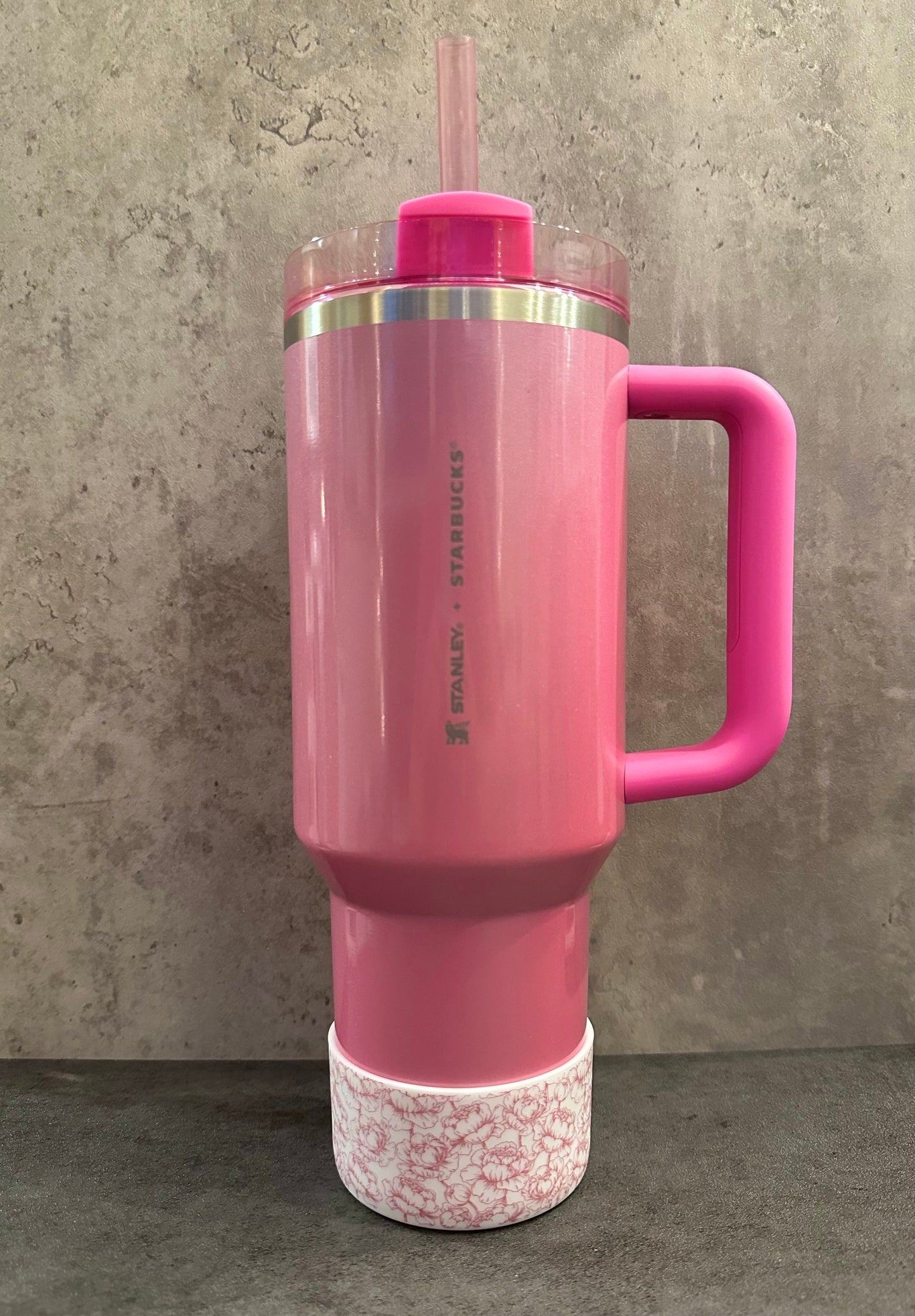 May include: A pink stainless steel tumbler with a black and white floral design on the bottom. The tumbler has a pink handle and a pink lid with a straw. The tumbler has the words 'STANLEY. STARBUCKS' printed on the side.