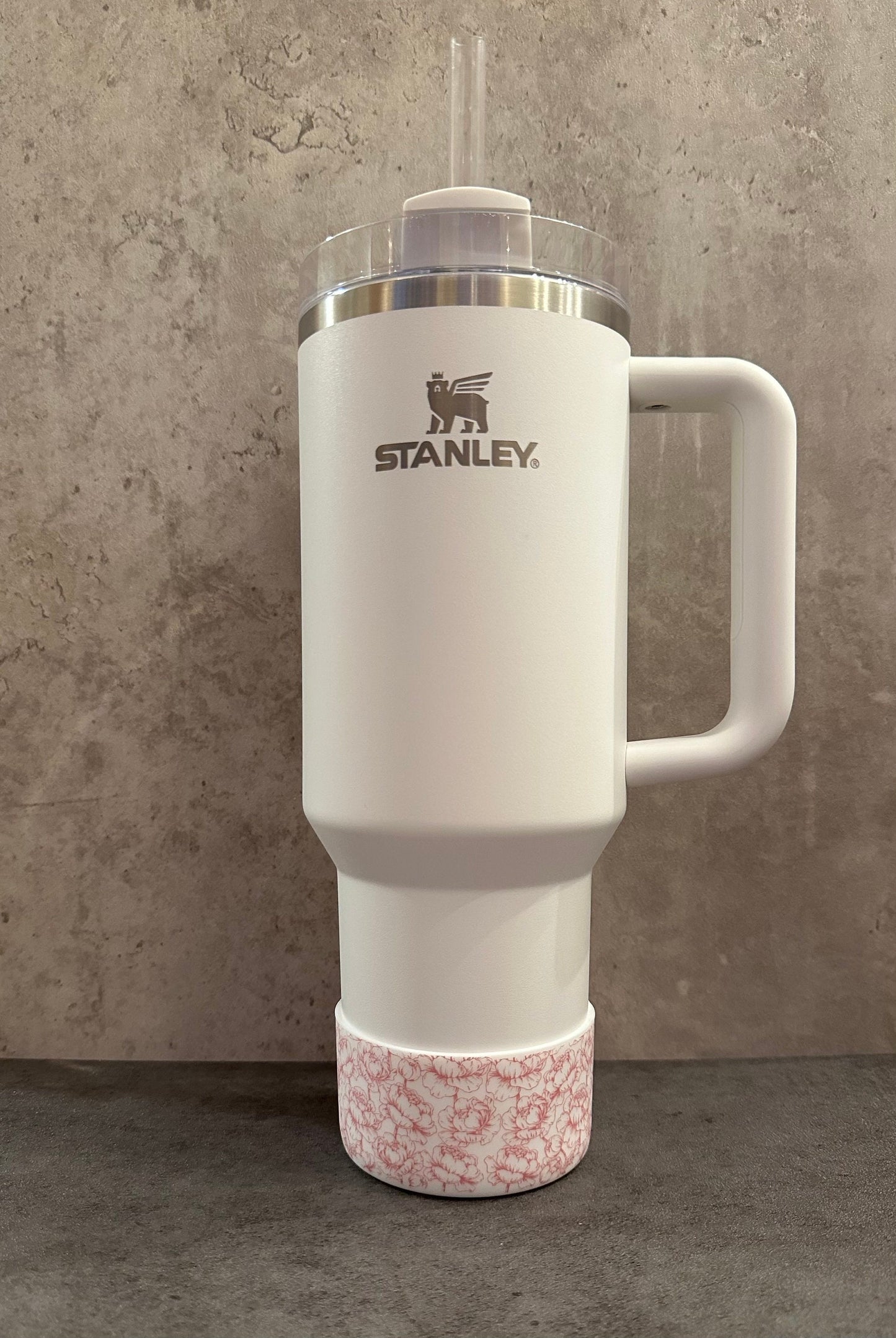 May include: White stainless steel travel mug with a black bear logo and the word 'STANLEY' printed on the side. The mug has a pink floral patterned base and a clear lid with a straw.