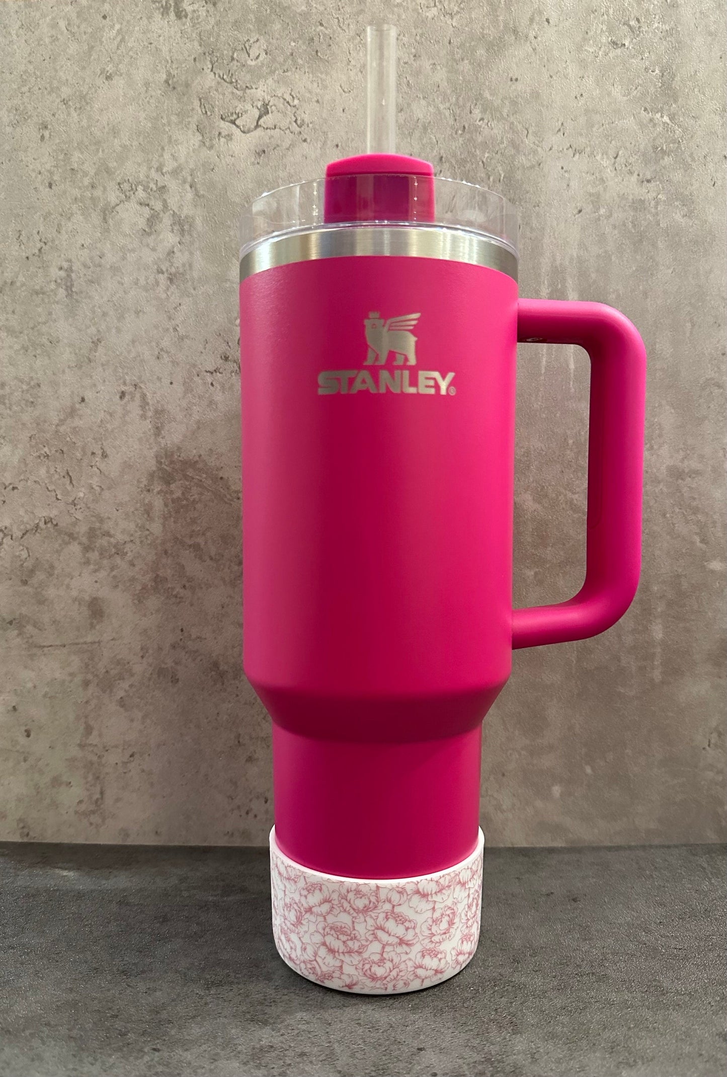 May include: A pink stainless steel travel mug with a black handle and a white lid with a straw. The mug has the Stanley logo on the front. The mug is sitting on a white and pink floral patterned base.