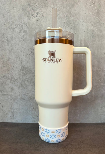 May include: A white stainless steel travel mug with a gold lid and handle. The mug has a gold logo that says &#39;STANLEY. SINCE 1913.&#39; The bottom of the mug has a blue and gold star of David pattern.