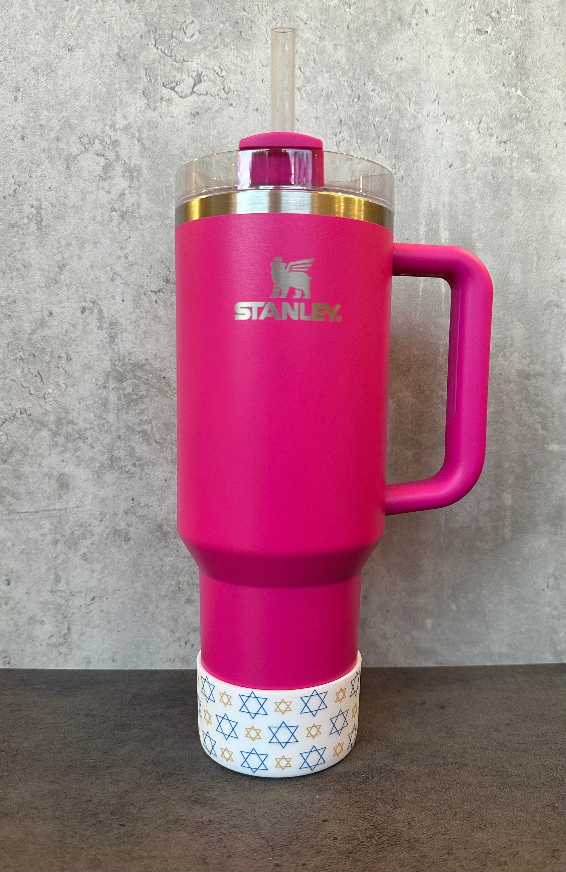 May include: A pink stainless steel travel mug with a handle and a straw. The mug has a white band with a pattern of gold and blue stars around the bottom. The mug has the word &#39;STANLEY&#39; printed on the side.