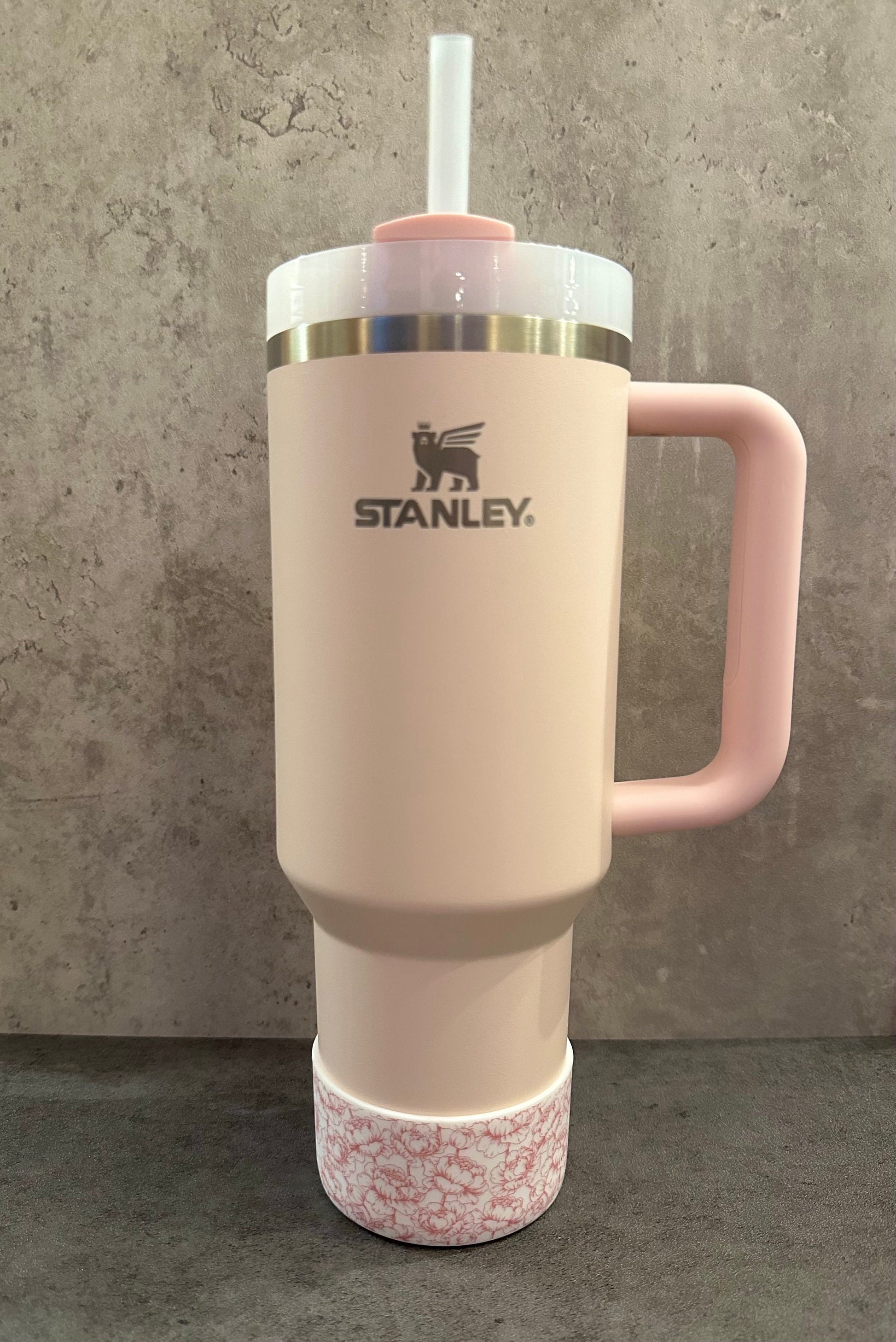 May include: A pink stainless steel travel mug with a handle and a straw. The mug has a floral pattern on the bottom and the Stanley logo on the side.