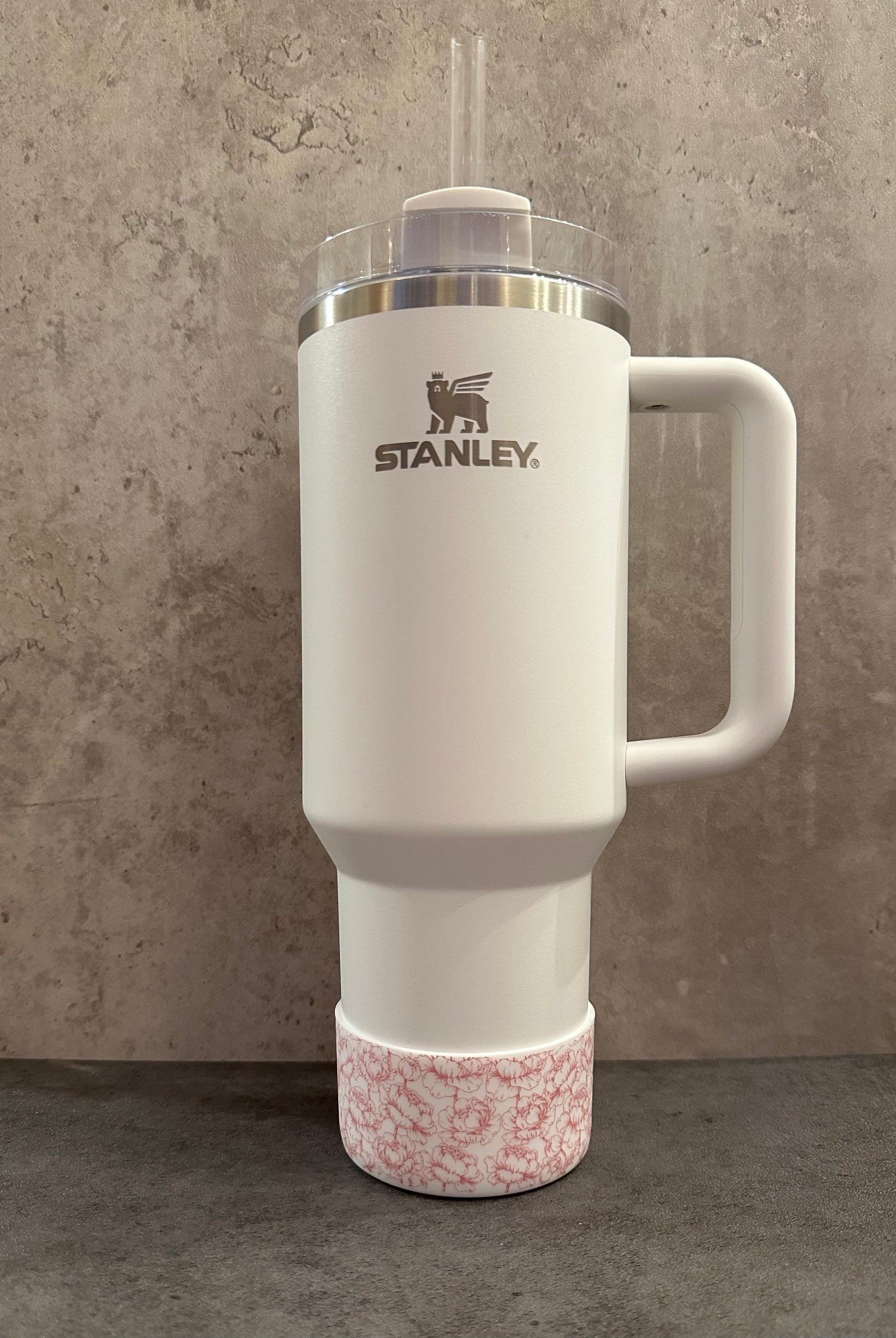 May include: White stainless steel travel mug with a black bear logo and the word &#39;STANLEY&#39; printed on the side. The mug has a pink floral patterned base and a clear lid with a straw.